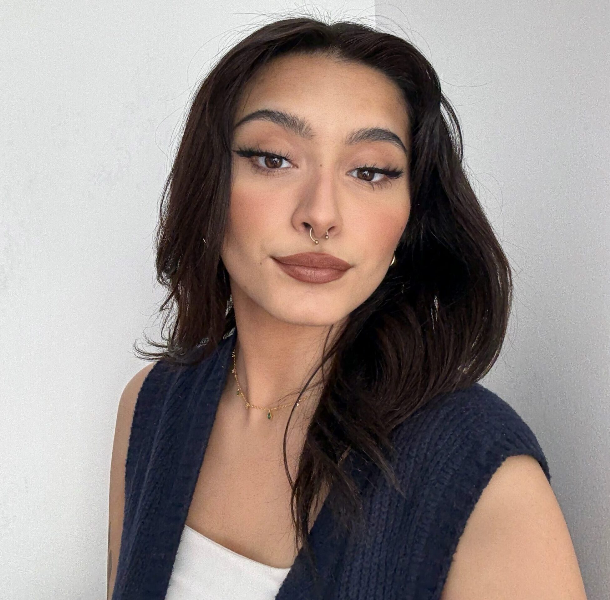 A person with long dark hair and winged eyeliner poses in front of a plain white wall, wearing a navy blue textured vest over a white top, a gold necklace, hoop earrings, and a septum piercing.