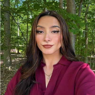 A person with long dark hair and makeup poses in front of a forest background, wearing a maroon v-neck blouse, a gold necklace, and two nose piercings.