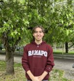 Sebastian in a maroon Ramapo College hoodie stands smiling with hands clasped in front, outdoors near a tree with green leaves and a grassy area.