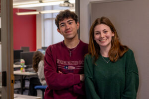 Two young adults, one wearing a maroon “Washington DC” sweatshirt and the other in a green sweater, stand smiling next to each other indoors. An office or classroom with desks and computers is visible in the background.