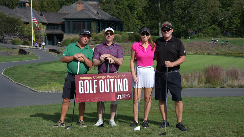 Four Golfers standing together on a lawn with a "Ramapo College Foundation Golf Outing" sign in front of them.