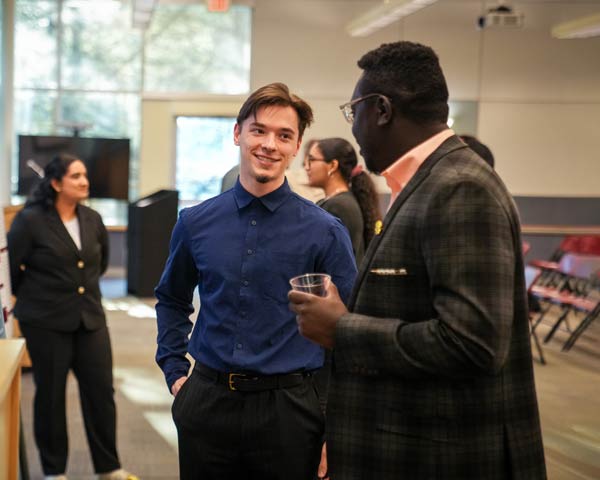 At Ramapo College's Center for Data, Mathematical, and Computational Sciences Fair event, two men chat warmly while holding drinks in a bright, modern room behind them.