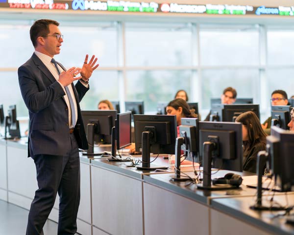 Charlie Lewis in a suit gestures while speaking with focused Ramapo College students at computer workstations in a bright, modern classroom.