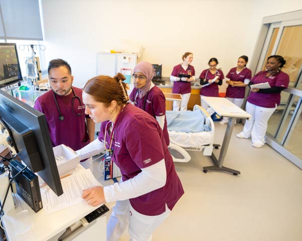 Six Ramapo College healthcare students in maroon scrubs are in a hospital room simulation lab. One works at a computer, while others observe and take notes. Medical equipment and a hospital bed are visible.