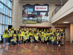 A large group of Ramapo College students in hard hats and high-visibility vests pose in a bright Veolia lobby beneath a wall display, conveying a cheerful site visit.
