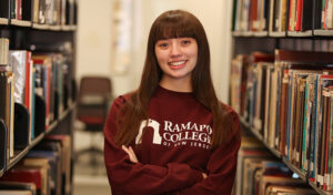 A young woman with long brown hair and bangs stands smiling in a library aisle, wearing a maroon Ramapo College of New Jersey sweatshirt. Shelves filled with books are visible on both sides.