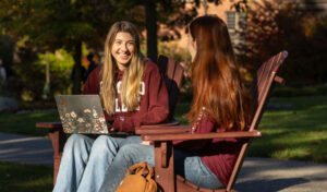 Two young women wearing maroon sweatshirts sit in Adirondack chairs outdoors, talking and smiling. One holds a laptop on her lap. There is a backpack on the ground and trees in the background.