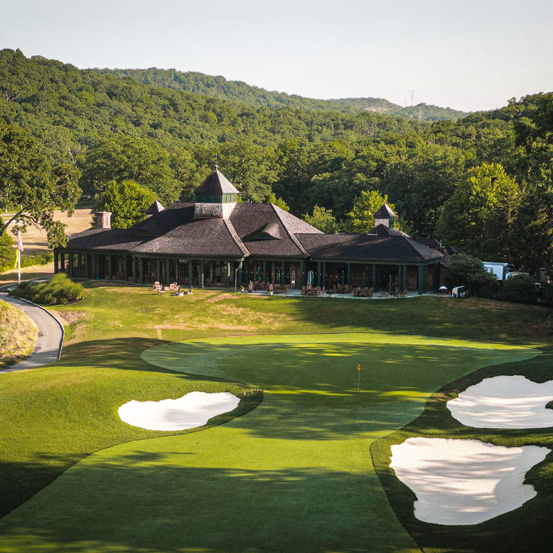 The Tuxedo Club with a large clubhouse with a dark roof sits surrounded by trees and hills, overlooking a manicured golf course green bordered by white sand bunkers under a clear sky.