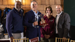 Dawkins, Pete, Jebb, and-Romano smiling beside banquet chairs as a man holds a trophy; the warmly lit wood-paneled setting conveys a celebratory award event.
