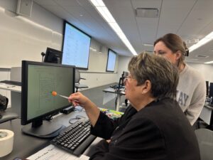 Two women in a classroom sit at a computer. One points at the monitor with a pencil while the other looks on. The room has multiple screens and rows of desks with computers.