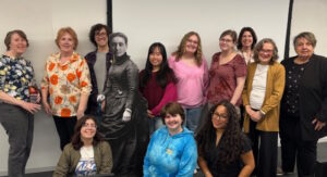 A group of twelve women pose and smile indoors, with two kneeling in front. They stand around a life-size black and white cutout of a woman in old-fashioned clothing. The background is a blank white wall.