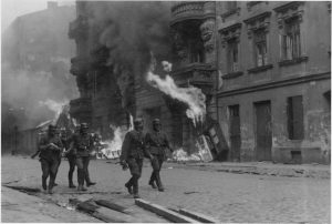 Black and white picture of Nazi soldiers wearing helmets walking through a burning Warsaw Ghetto