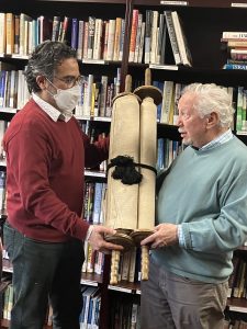 Rabbi Norman Patz with white hear and a goatie hands a Torah, bound with a black chord, to Jacob Labendz, in a red sweater and COVID mask