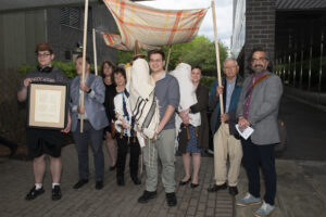 Students and dignitaries standing outside on Ramapo campus, holding three Torahs and a framed certificate.