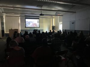 A large room with about 70 people watching something being projected onto a screen at the front of the room.