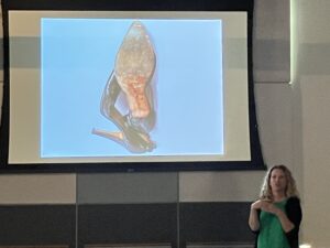 Woman with curly blond hair in a green shirt and black jacket standing to the side of a projection screen with a picture of high-heeled shoes on it.