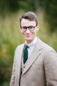 David Austin Walsh. A white man with brown hair and glasses, wearing a beige suit and a green tie.