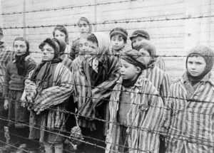Children wearing adult-sized, striped prisoner uniforms, standing behind barbed wire.