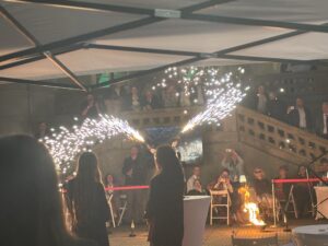 Picture of a woman twirling sparkling fire taken from behind in front of an audience standing on double stairs outside of a building.