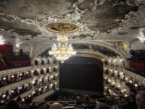 Interior of a grand opera house with three levels of lodges, a baroque ceiling with a chandelier, and a red curtain drawn in front of the stage