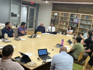 People sitting around a large, rectangular wooden table. Some have laptops.