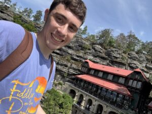 Smiling young man in a blue tee-shirt with an historic hotel, built into a rock wall, in the background.