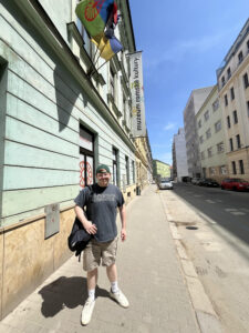 Young man in a gray shirt and cap with a shoulder bag, standing in front of a green building with a Romani Flag.