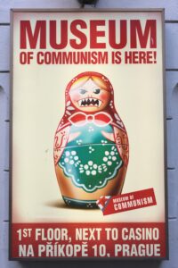 Poster for the Museum of Communism in Prague. It is off-white with red lettering: "Museum of Communism is Here!" It includes the institutions old address. The picture is of a matryoshka with fangs. 