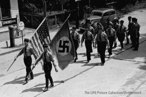 Picture of men marching in uniform on a street. At the head are two standard bearers. One caries the Nazi flag and the other the US flag.
