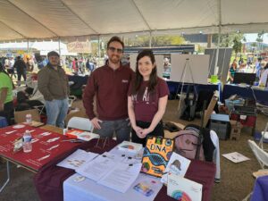 Two people stand behind a table at an outdoor event booth with science materials, including a DNA book, flyers, and posters. Other booths and attendees are visible under a large tent in the background.