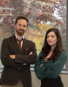 Two people standing indoors with arms crossed, posing confidently in front of a large window. Outside, autumn trees with red and orange leaves are visible. The man wears a plaid blazer and tie; the woman wears a green blouse.