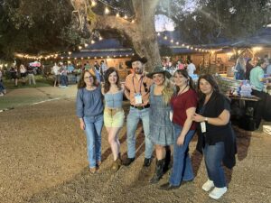 Six smiling people pose together outdoors at a nighttime event under string lights, with a large tree, rustic buildings, and other attendees in the background. Most are casually dressed and some wear cowboy hats and boots.