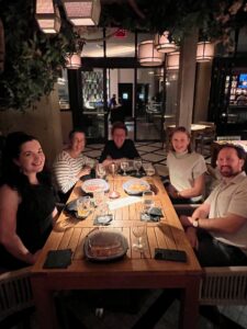 Five people sit smiling around a wooden table at a dimly lit restaurant, with drinks and food on the table. The atmosphere is cozy, with warm lighting, greenery above, and stylish decor in the background.