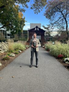A man in a gray blazer and dark pants stands on a garden path holding a book, with tall grasses and flowers on either side and a wooden gazebo structure and glass building in the background. Trees show some autumn colors.