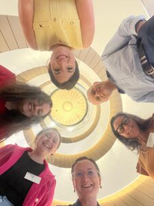 Six people stand in a circle, smiling and looking down at the camera beneath them. Above them is a spiral staircase with a bright light at the center, creating a circular pattern overhead.