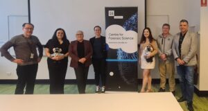 Seven people stand indoors beside a Centre for Forensic Science banner; some are holding koala toys. The setting appears to be a conference or seminar room with a green floor and white walls.