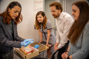 A woman in gloves explains lab materials to three people gathered around a table, all attentively watching. A handwritten note with figures is visible on the wall in the background.