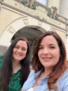 Two women smiling in front of the entrance to Taronga Zoological Park, with the ornate building sign visible above them.