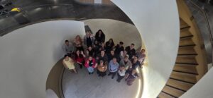 A group of people stands together, looking up at the camera, inside a modern building next to a curved staircase and white circular wall.
