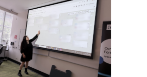 A person in a black dress points at a large projected screen displaying a web browser with blurred content in a classroom or seminar setting.