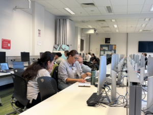 Several people work at computers in a bright classroom; two women in the foreground collaborate over notes, while others focus on their screens.