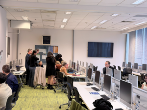 People are working and talking in a modern computer lab with many desktop computers. Some are seated at desks while a small group stands and talks near the center. Large windows let in natural light.