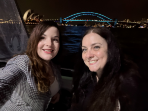 Two women smiling for a selfie at night with the Sydney Opera House and Sydney Harbour Bridge, both lit up, in the background across the water.