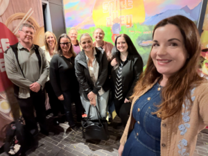 A group of eight adults smile while posing for a selfie on a cobblestone street at night, with a colorful Smile Alley mural in the background.