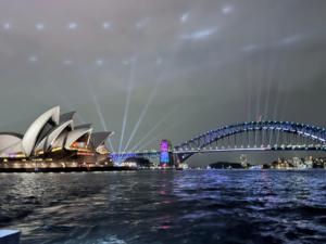 Sydney Opera House and Harbour Bridge at night, illuminated with colorful lights and beams reflecting on the water under a cloudy sky.