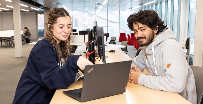 A woman and a man sit at a desk in a modern, brightly lit office space, looking at a laptop together. The woman is pointing at the screen while the man watches attentively.