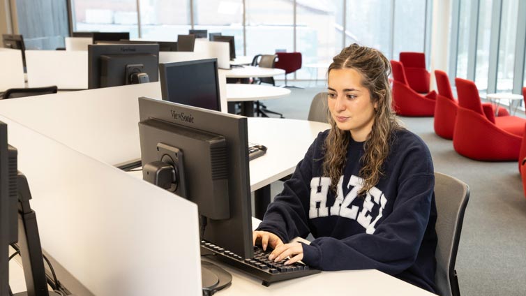 A young woman with long brown hair, wearing a navy blue HAZE sweatshirt, sits at a desk in a modern computer lab, working on a desktop computer. Red chairs and large windows are visible in the background.