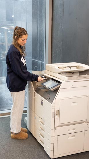 A woman wearing a dark sweatshirt, light pants, and slippers is using a large photocopier in an office space with large windows.