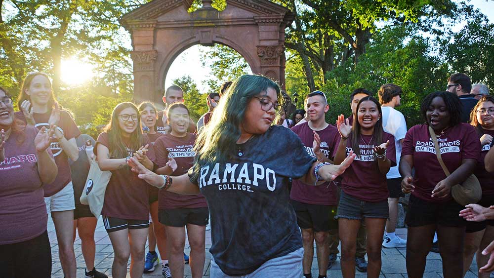 Diverse group of Ramapo College Students as the sun is setting by the Arch with a student dancing in the forefront