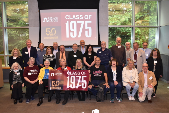 Group shot of 24 former students from the class of 1975, holding a banner indoors